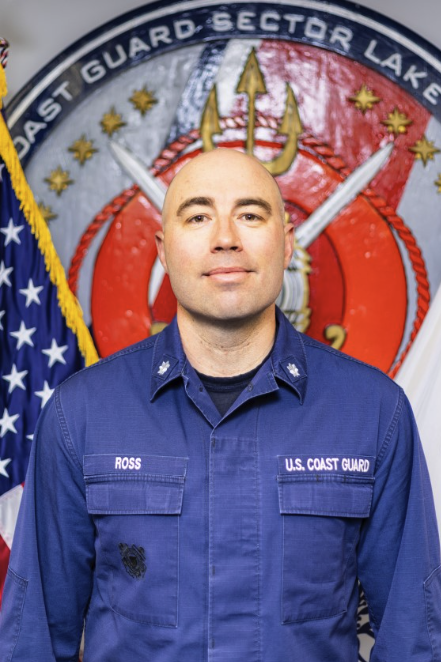 U.S. Coast Guard officer in uniform standing before a Sector emblem and U.S. flag